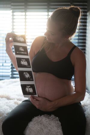 Pretty pregnant woman holding ultrasound photos while sitting on bed and relaxing at home. Maternity, pregnancy, new life and family planning conceptの写真素材