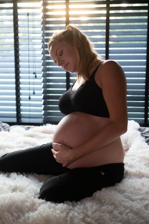 Pretty pregnant woman in black leggins and bra suffering lower back pain while sitting on her bed at home. Pregnancy, maternity, health care, gynecology, medicineの写真素材
