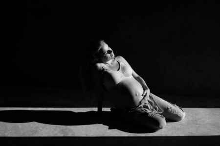 Pretty pregnant woman in denim jumpsuit and bra sitting on the concrete floor in sunlight. Maternity conceptの写真素材