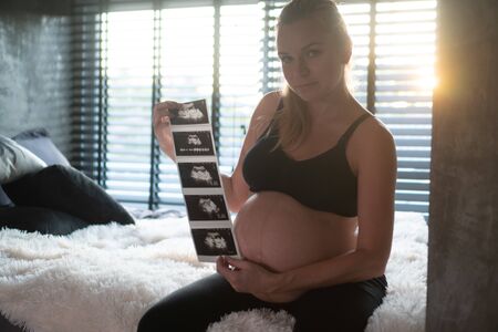 Pretty pregnant woman holding ultrasound photos while sitting on bed and relaxing at home. Maternity, pregnancy, new life and family planning conceptの写真素材