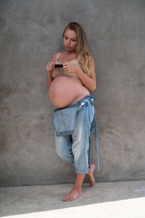 Pretty pregnant woman in denim jumpsuit and bra drinking coffee while standing over gray wall background. Maternity, pregnancy, new life healthy food conceptの写真素材