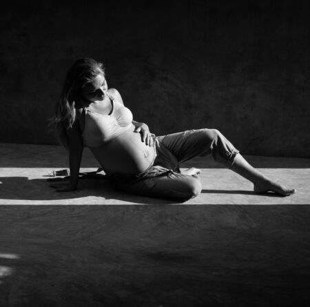 Pretty pregnant woman in denim jumpsuit and bra sitting on the concrete floor in sunlight. Maternity conceptの写真素材