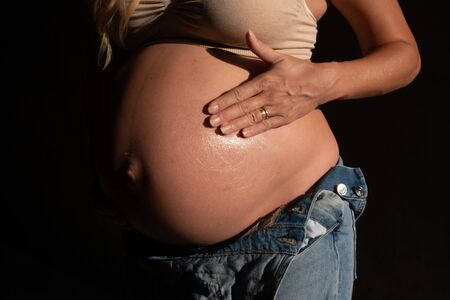 Cropped image of pregnant woman in denim jumpsuit and bra applying lotion on her belly. Maternity, pregnancy, new life and health care conceptの写真素材