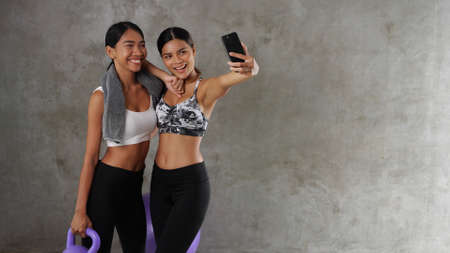 Two beautiful young Asian women doing selfie photo with smart phone at the gym. Happy female friends spending their free time at the sport centreの写真素材