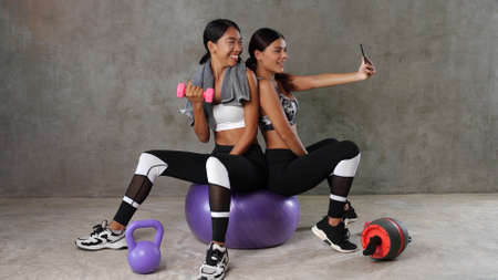 Two beautiful young Asian women posing with fitness ball and dumbbell while doing selfie photo at the gym. Happy female friends spending their free time at the sport centreの写真素材