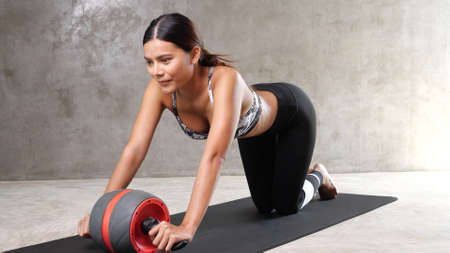 Beautiful young Asian woman exercising with ab wheel on the yoga mat at the gym. Happy girl spending her free time at the sport centreの写真素材