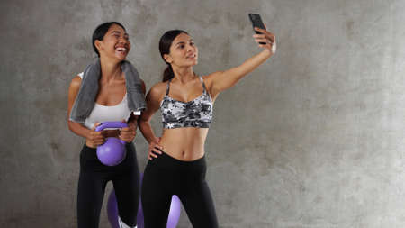 Two beautiful young Asian women doing selfie photo with smart phone at the gym. Happy female friends spending their free time at the sport centreの写真素材