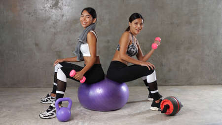Two beautiful young Asian women posing with fitness ball and dumbbell at the gym. Happy female friends spending their free time at the sport centreの写真素材