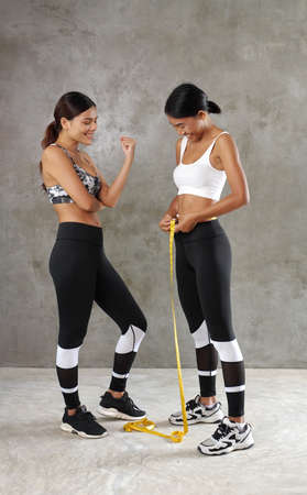 Two beautiful young Asian women measuring their waist at the gym. Happy female friends spending their free time at the sport centreの写真素材