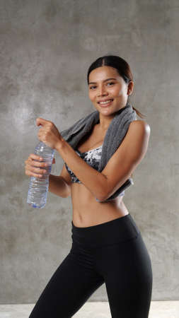 Beautiful young Asian woman posing with water and towel at the gym. Happy girl spending her free time at the sport centreの写真素材