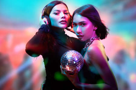Two young flirting cute Asian women in black bodysuits with headphones and small disco ball posing...