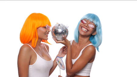 Two young smiling Asian women in bright color wigs and sunglasses posing with photo booth props and small disco ball over white background. Party, photo booth, friendship, fun time conceptの写真素材