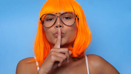 Portrait of beautiful happy Asian woman in bright orange wig and glasses posing over blue backgroundの写真素材