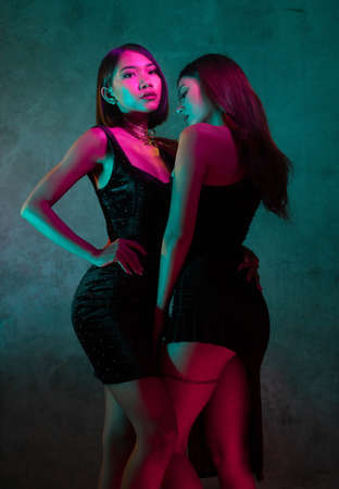 Portrait of two young cute Asian women in elegant dresses on the pink and yellow neon lightsの写真素材