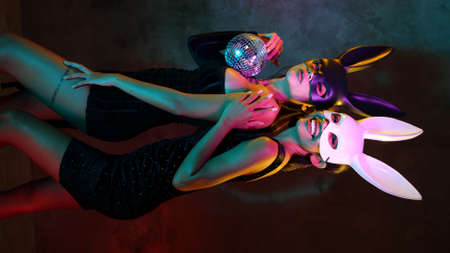 Portrait of two young flirting cute Asian women in black and white rabbit mask playfully posing for camera with small disco ball on the neon lightsの写真素材