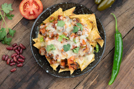A plate of delicious corn chips nachos in cheddar cheese with beans, tomato, green chili, black olives, cilantro served with guacamole, sour cream and tomato salsa dip on the side isolated on the wooden tableの写真素材