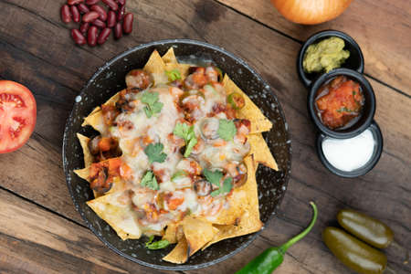 A plate of delicious corn chips nachos in cheddar cheese with beans, tomato, green chili, black olives, cilantro served with guacamole, sour cream and tomato salsa dip on the side isolated on the wooden tableの写真素材
