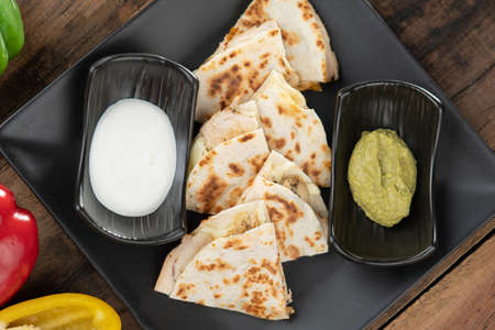 A toasted tortilla with chicken, peppers, red onion and mozzarella served with a guacamole dip and sour cream isolated on wooden tableの写真素材