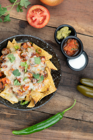 A plate of delicious corn chips nachos in cheddar cheese with beans, tomato, green chili, black olives, cilantro served with guacamole, sour cream and tomato salsa dip on the side isolated on the wooden tableの写真素材