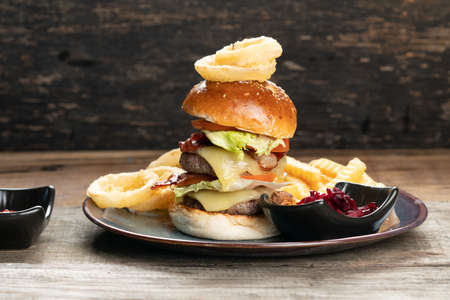 Big burger with beef, bacon, tomato, cheese, lettuce and onion served with french fries, onion rings and beetroot salad on the side isolated on rustic wooden tableの写真素材