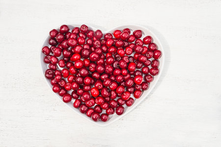 Cranberries in heart shaped bowl on white wooden background, top viewの写真素材