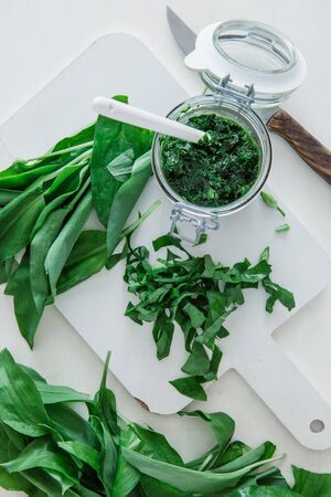 A glass of pesto  and green leaves of wild garlic with kitchen utensils on a white table, top view