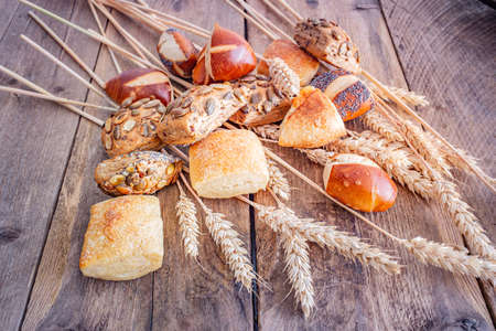 A large selection of freshly baked rolls on a wooden, rustic background with a bundle of wheat