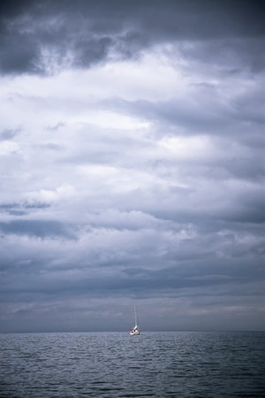 Lonely boat sails on the calm sea beneath a stormy sky. Light is breaking through the cloudsの写真素材