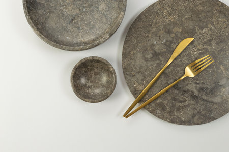 Top view of brown marble tray, plate, bowl and gold cutlery on white background. Food presentation,...