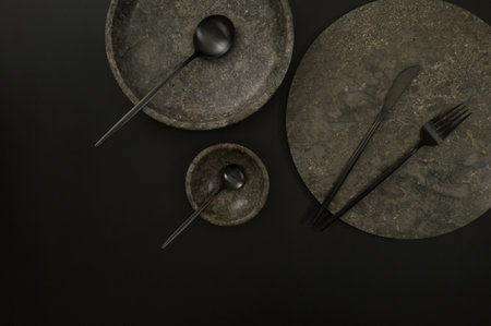 Top view of brown marble tray, plate, bowl and black cutlery on black background. Food presentation,...