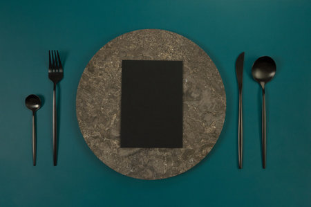 Top view of brown marble plate, black cutlery and black card on dark green background. Food presentation, stone tray. Table setting, copy space, flat lay.の写真素材