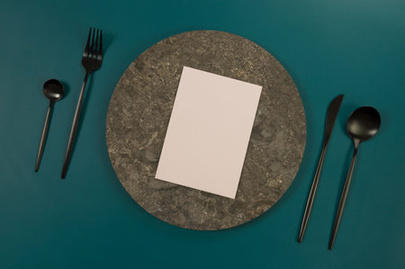 Top view of brown marble plate, black cutlery and pink card on dark green background. Food presentation, stone tray. Table setting, copy space, flat lay.の写真素材