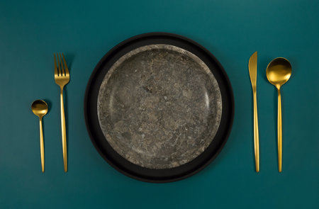 Top view of brown marble plate, black plate, gold cutlery on dark green background. Food presentation, stone tray. Table setting, copy space, flat lay.の写真素材