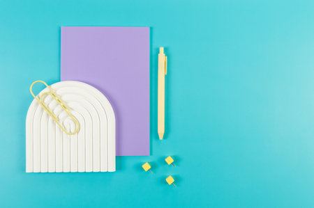 Top view of purple notebook, yellow pen, paperclip, pins, white arch tray on blue background. School, office wallpaper. Flat lay, copy space.の写真素材