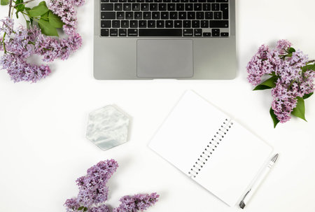 Top view of purple lilac flowers on white background. Laptop computer, white notebook, pen and...