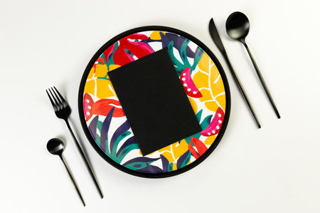 Top view of black tray, colorful plate, black cutlery on white background. Black card, exotic plant...
