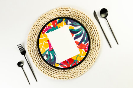 Top view of brown wicker table mat, colorful plate, black cutlery on white background. Black tray,...