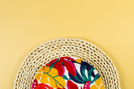 Top view of brown wicker table mat, colorful plate on yellow background. Straw round tray, exotic plant plate, summer picnic. Flat lay, copy space.の写真素材