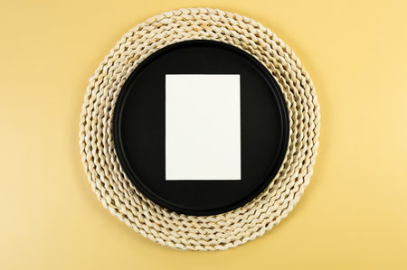 Top view of brown wicker table mat, black plate, white card on yellow background. Straw round tray, flat lay, copy space.の写真素材