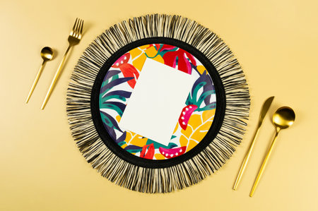 Top view of black wicker table mat, colorful plate, gold cutlery, white card on yellow background. Straw round tray, exotic plant plate, summer picnic. Flat lay, copy space.の写真素材