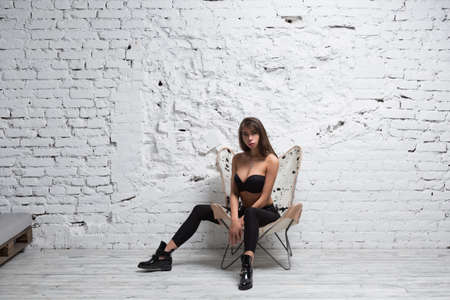Seductive brunette woman posing on a modern chair against white grungy brick wallの写真素材