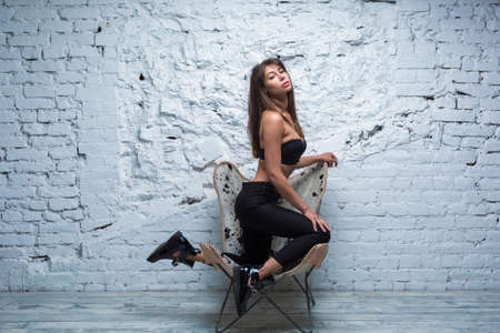 Seductive brunette woman posing on a modern chair against white grungy brick wallの写真素材
