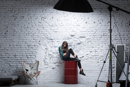 Stylish brunette woman sitting on a red barrel against white grungy brick wall. Professional photo studio with modern lighting equipmentの写真素材