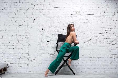 Topless brunette woman sitting on a directors chair against white grungy brick wall.の写真素材