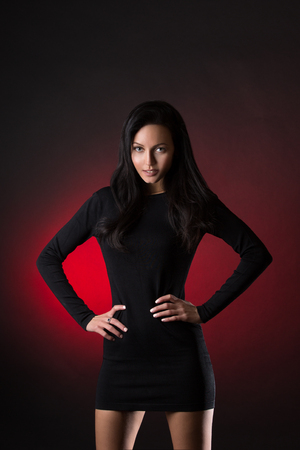 Beautiful brunette woman posing over dark red background in short black dressの写真素材