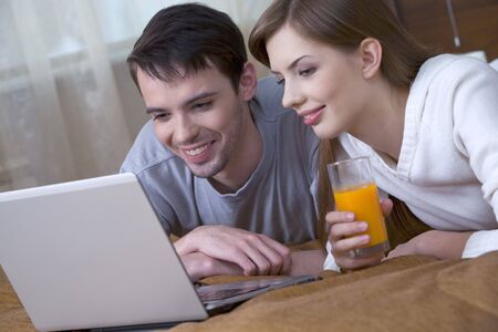 The young man and the girl with glass of juice in a hand, resting together, look at the laptop screenの写真素材