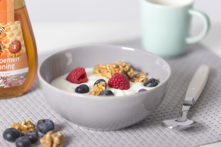 Bowl of yogurt with fresh raspberries, blueberries and nuts with spoon on gray with green cup and honey at the side. Brekfastの写真素材