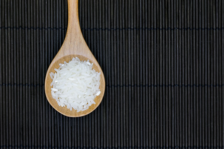 uncooked jasmine rice in wooden spoon on the black bamboo mat backgroundの写真素材