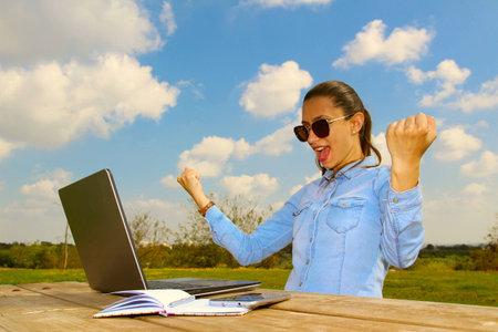 A happy woman with a laptop sitting in the garden, with arms up in the summerの写真素材