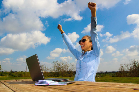 A happy woman with a laptop sitting in the garden, with arms up in the summerの写真素材